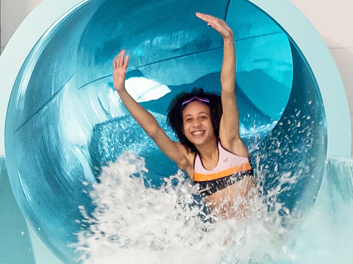 A child emerging smiling from the end of a blue tube flume into a splash of water