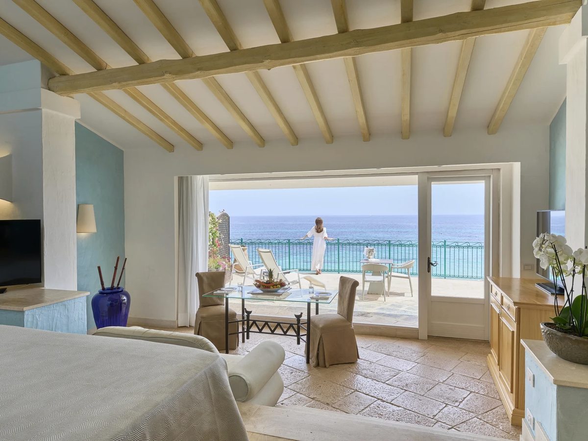 A room with a beamed ceiling and wide doors opening onto a terrace with loungers directly above the sea