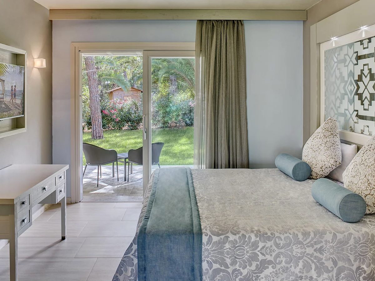 A double bedroom with sliding doors onto a paved terrace with table and chairs, set in a green garden