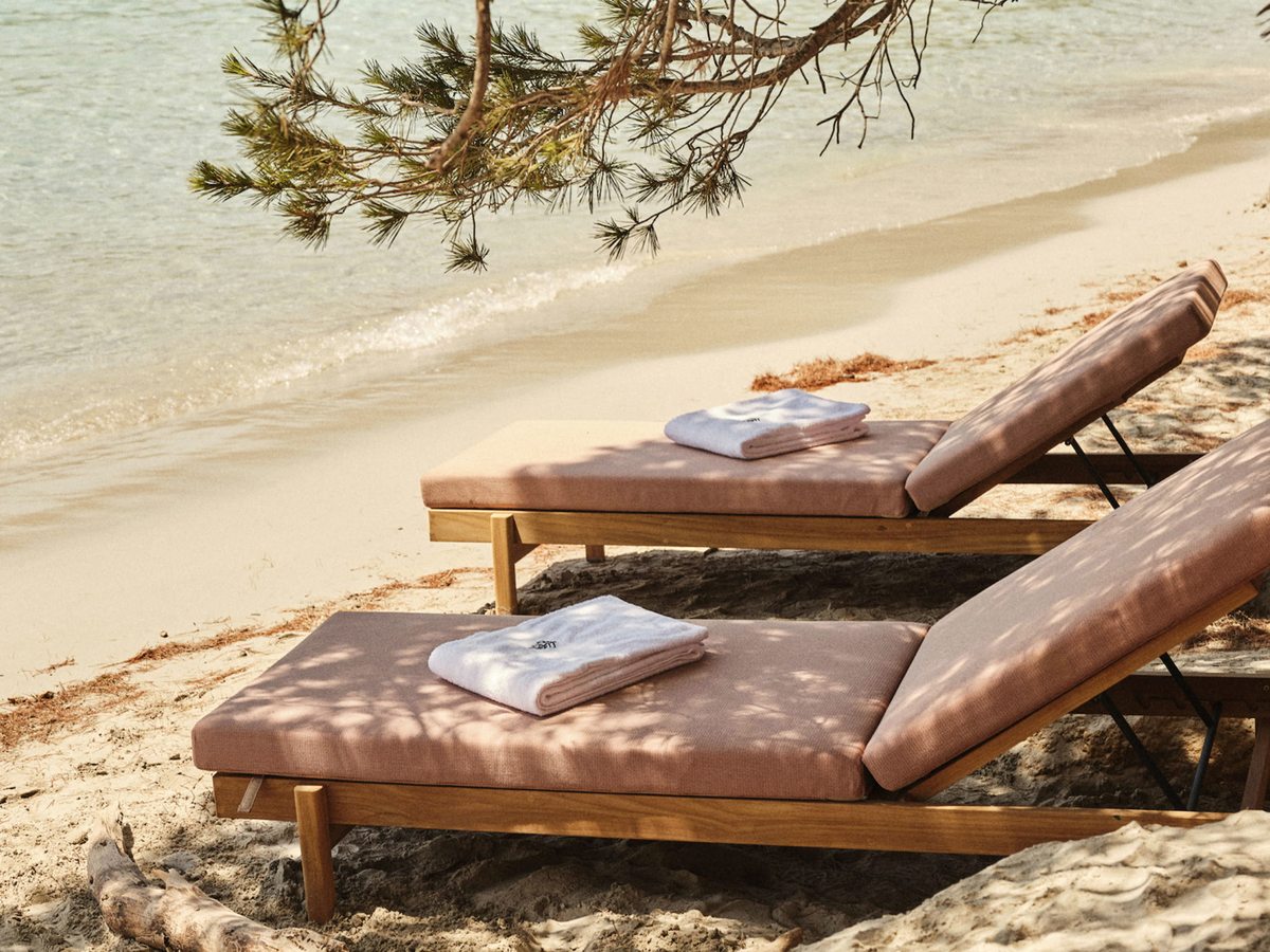 Two thickly cushioned wooden sun loungers with folded towels on the sand under pine shade, at the edge of the beach.