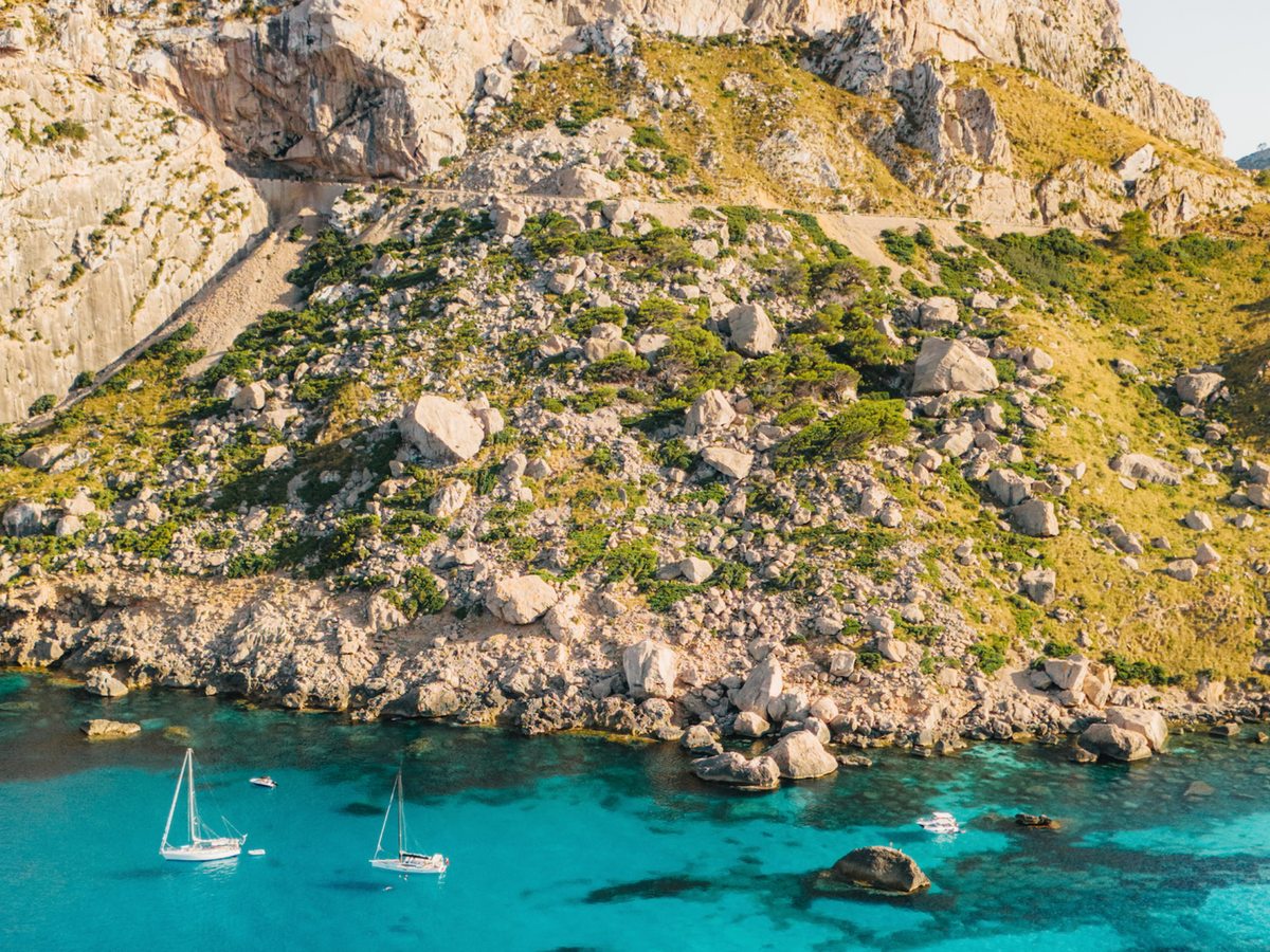 Aerial view of a rocky, pine-covered headland enclosing a cove of clear turquoise water with a few small boats.