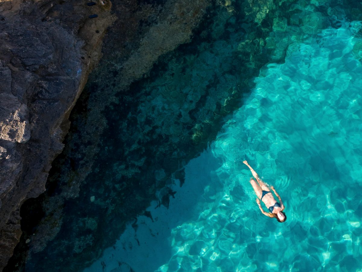 Overhead view of a snorkeller floating in clear turquoise water beside dark rocks in a cove.