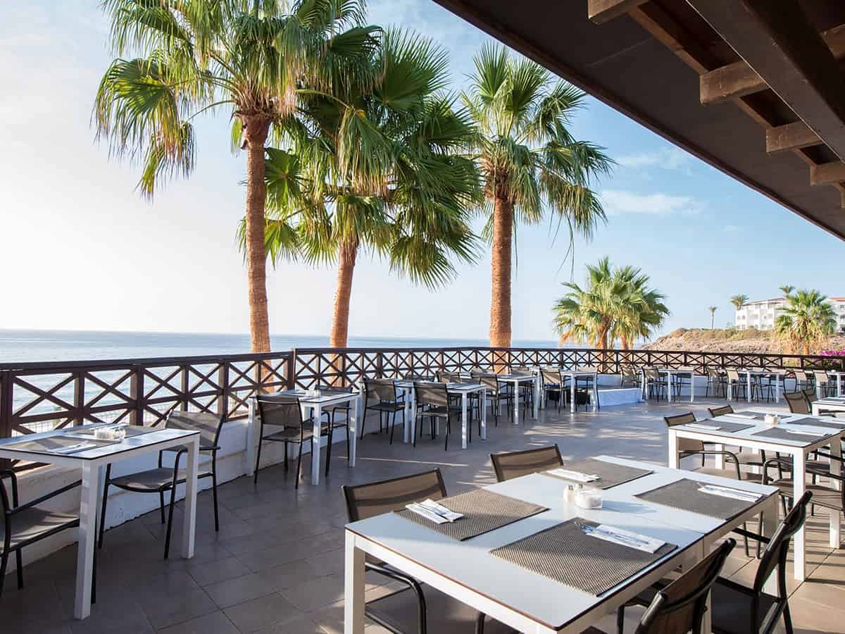 An open-air terrace restaurant with white-clothed tables under a timber pergola, tall palms and the sea beyond.