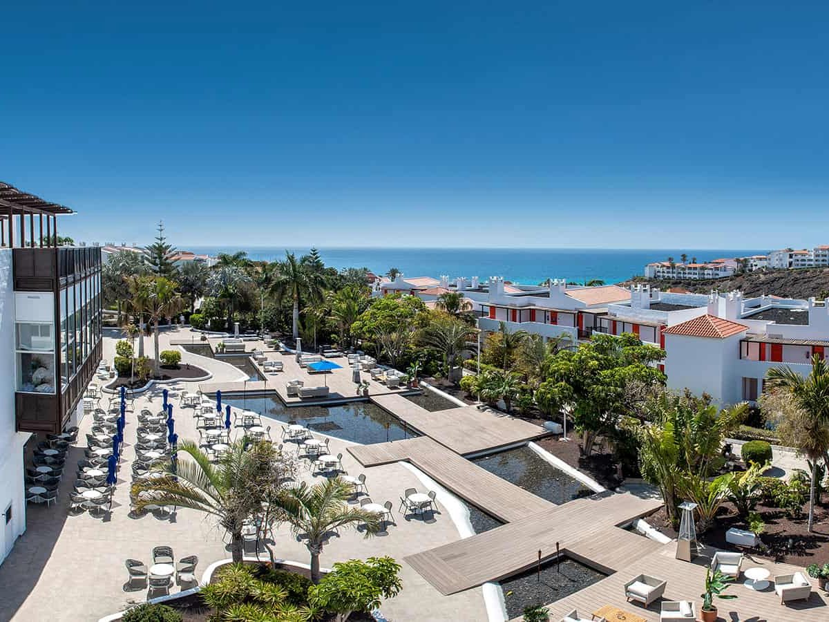 A landscaped central courtyard of palms and planting with dark ornamental water channels and a terrace of dining tables, the sea on the horizon.