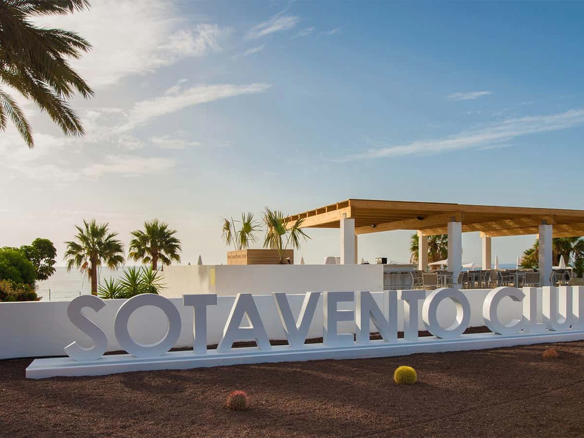 Large white free-standing letters spelling Sotavento Club on gravel, a timber pergola terrace and palms behind and the sea beyond.