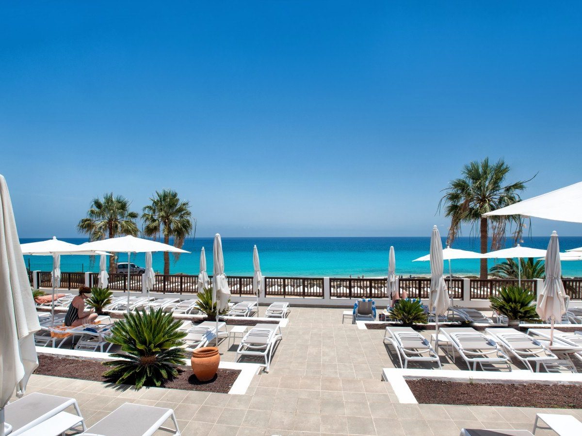 Rows of sun loungers under white umbrellas on a paved terrace at the edge of the beach, the sea beyond.