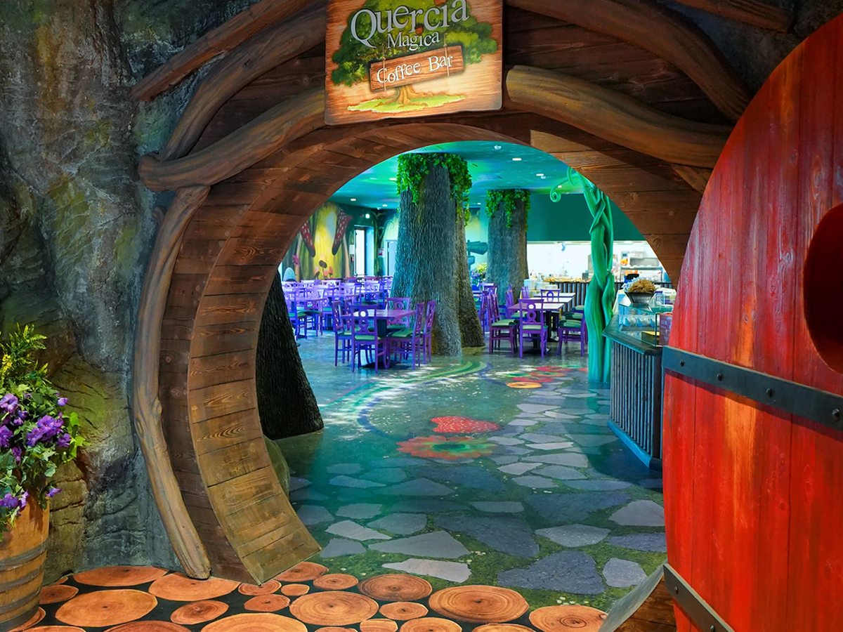 The Quercia Magica Coffee Bar entrance, a round wooden door set in a tree-trunk archway leading into a woodland-themed dining room with purple chairs.