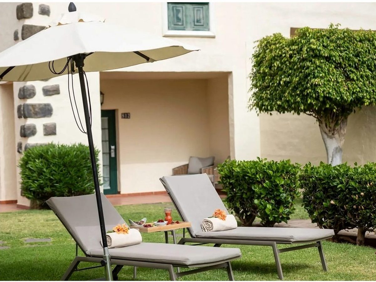 Two cushioned sun loungers and a parasol on the lawn of a ground-floor unit's private garden, in front of a cream villa with a green-shuttered door.