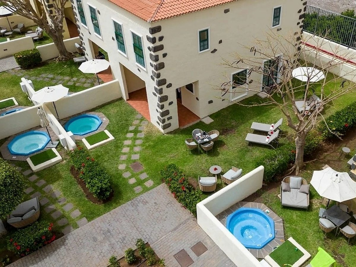 An aerial view of walled private gardens beside a cream villa with volcanic-stone trim, each garden with a round sunken jacuzzi, lawn, paving and seating.