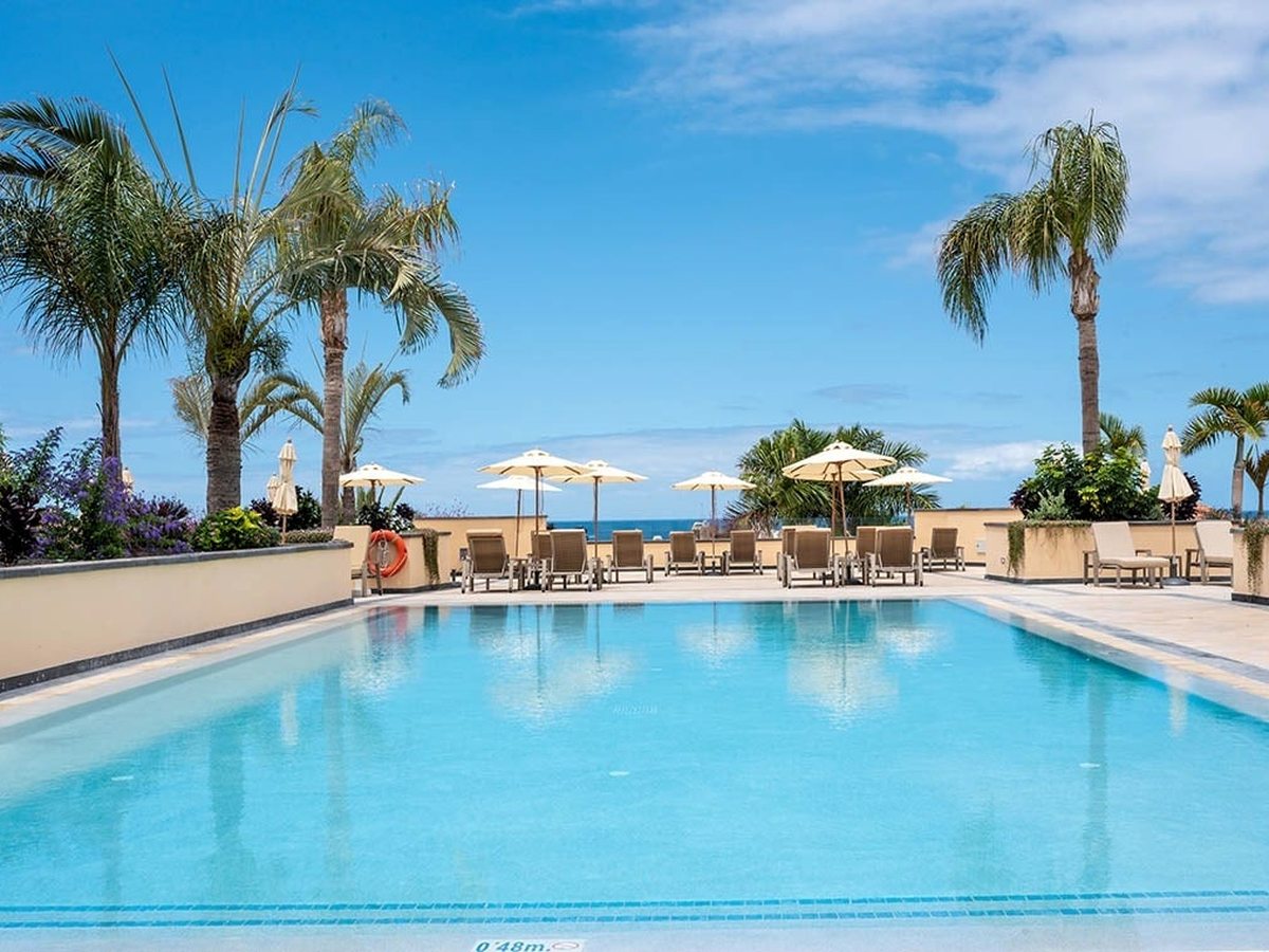 A pool with a broad, gently sloping shallow end marked at 0.48 metres, flanked by palms, with white parasols and the sea on the horizon.