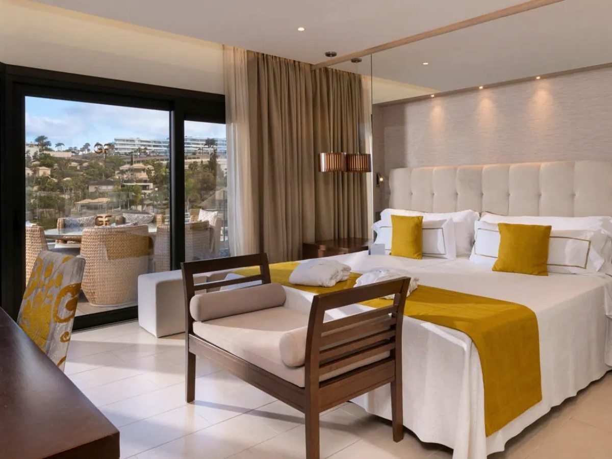 A double bed with a mustard runner and cushions beside sliding glass doors onto a furnished balcony.
