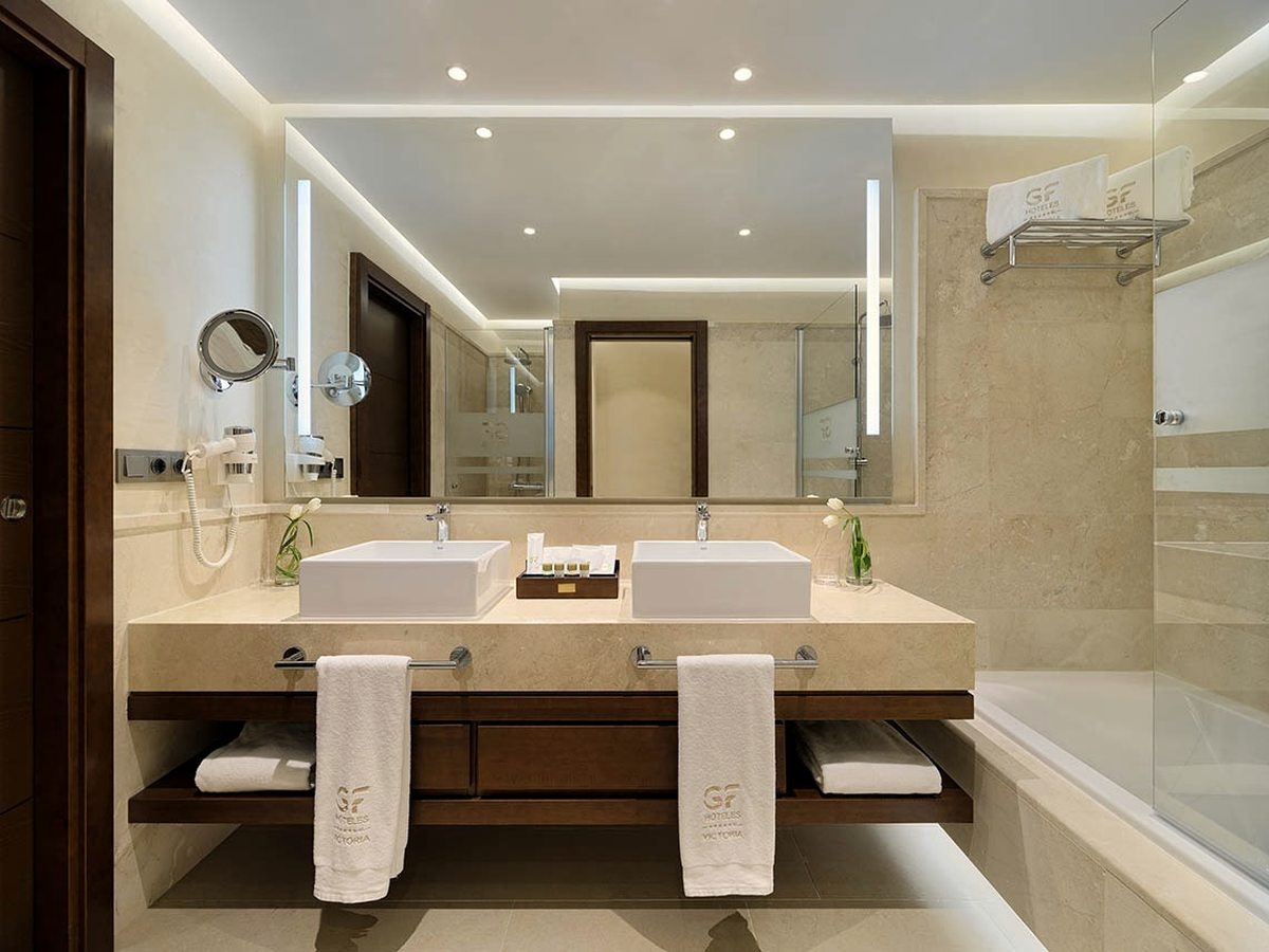 A marble bathroom with twin square vessel basins, a large lit mirror and a bath behind a glass screen.