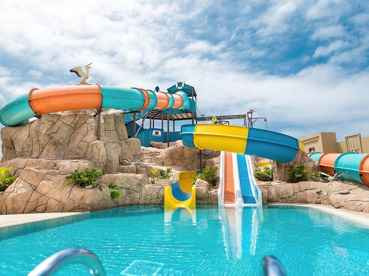 Colourful blue, yellow and orange water slides winding over a moulded rock structure into a shallow pool under a bright sky.