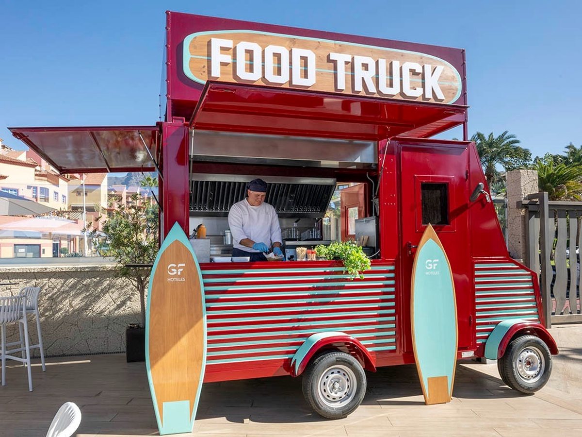 A red vintage food truck marked Food Truck with a chef cooking inside and turquoise surfboards propped against it.