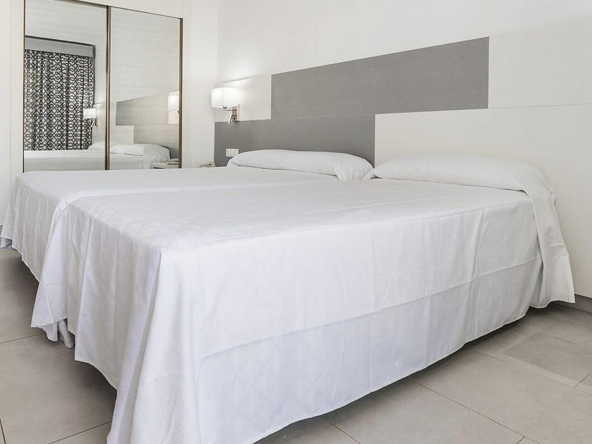 A double room with a large bed under white bedding against a grey wall panel, with a mirror and a tiled floor.