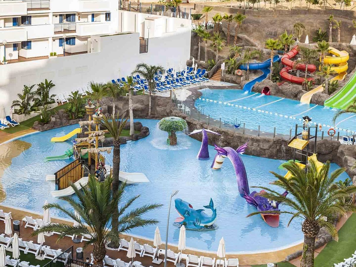 An aerial view of the children's water park with a pirate ship, a purple sea-dragon feature, tube slides and a rocky backdrop, the Los Patos Park name on the pool floor.