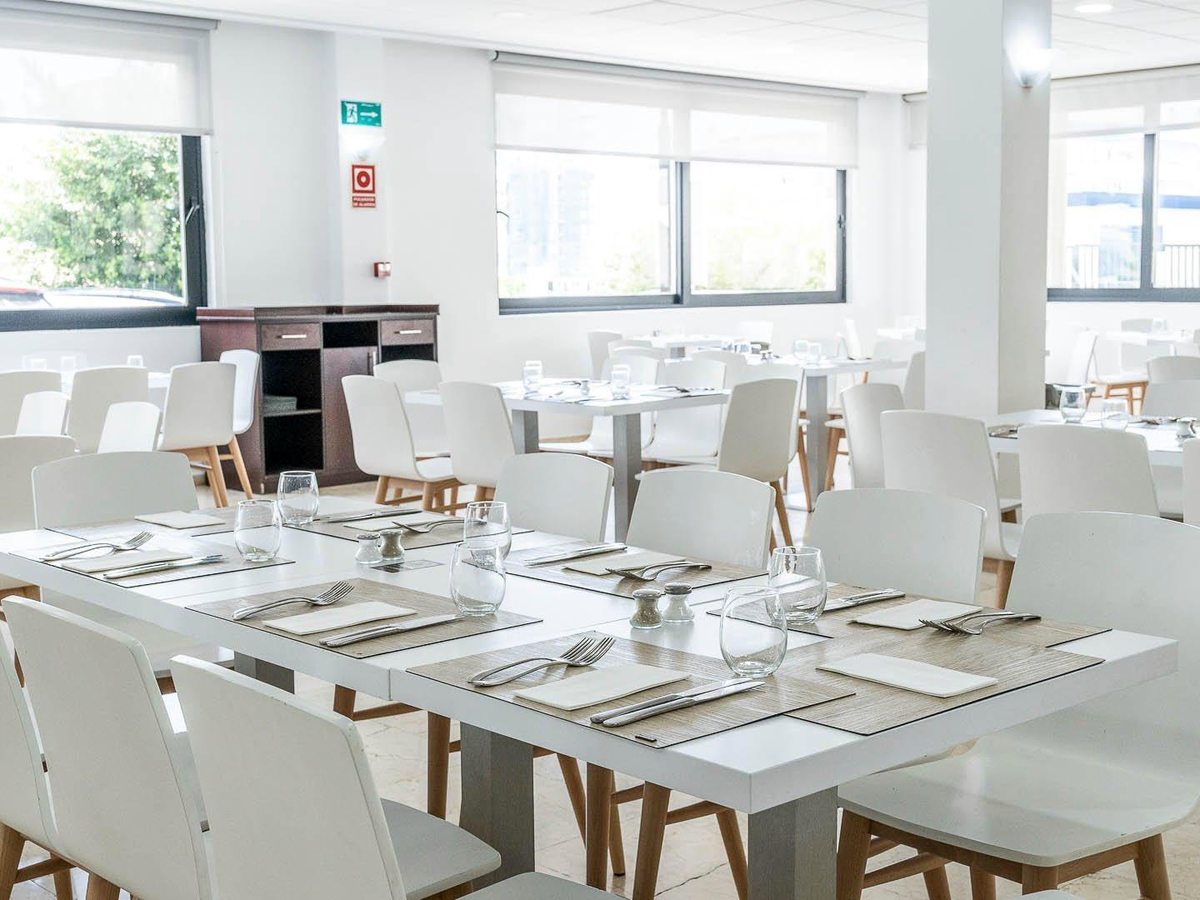 A bright buffet dining room with white chairs and wood-topped tables set with glasses and place mats, windows along one side.