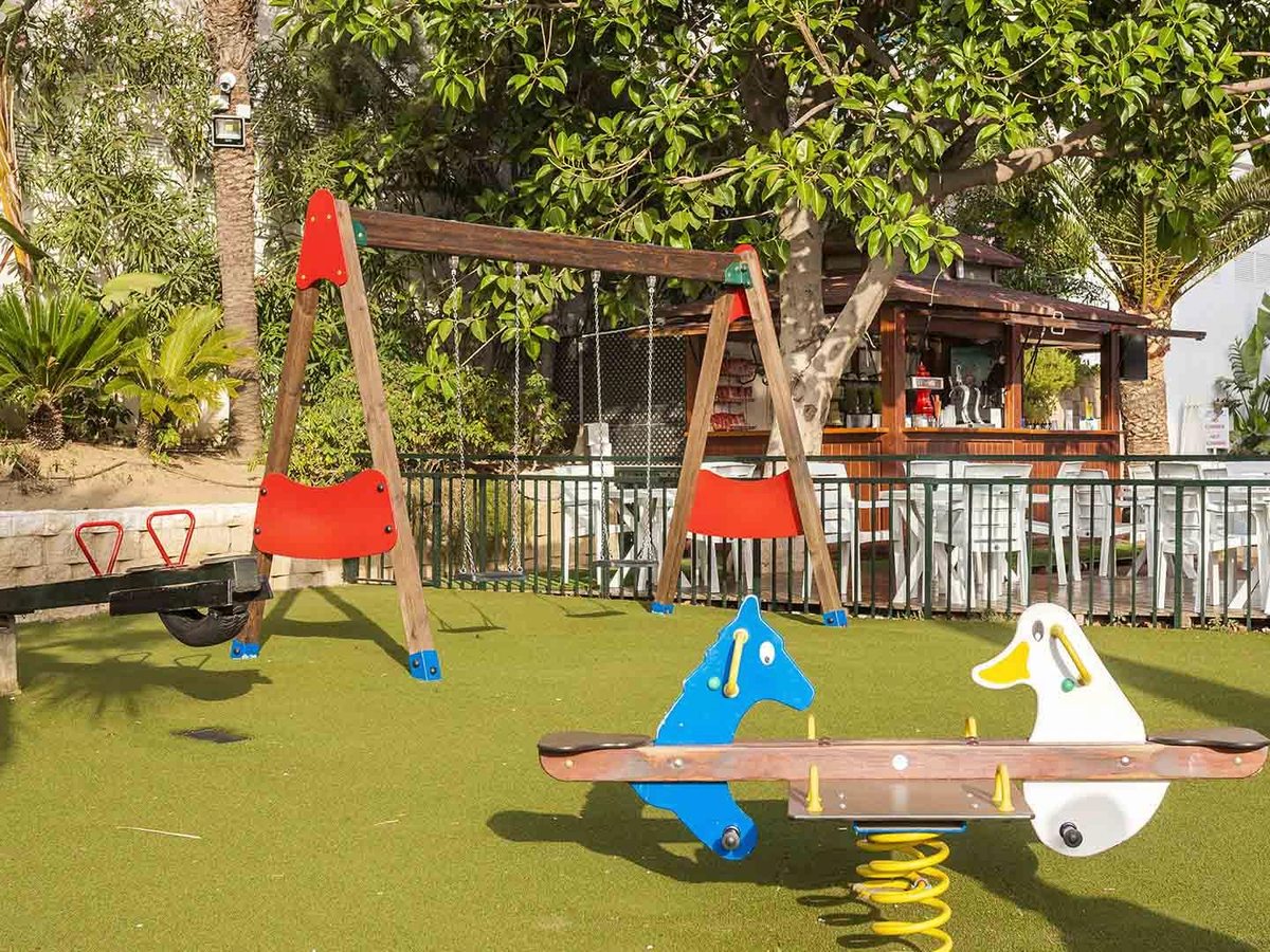 An outdoor playground with red swings and spring-mounted ride-on animals on artificial grass, with a bar building behind.