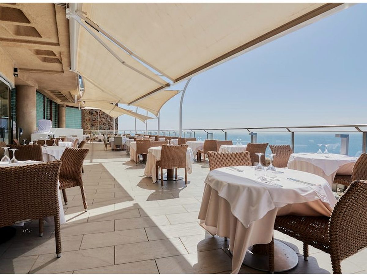 Outdoor restaurant terrace with white-clothed tables and wicker chairs under awnings, beside a glass rail above the sea.
