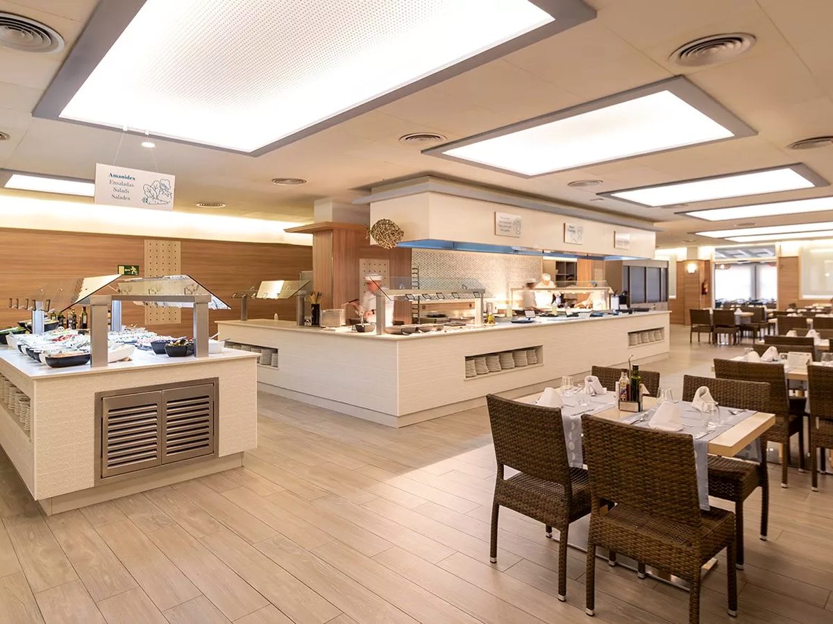 The buffet restaurant with a central serving island of hot dishes under bright ceiling panels, and wicker chairs at set tables.