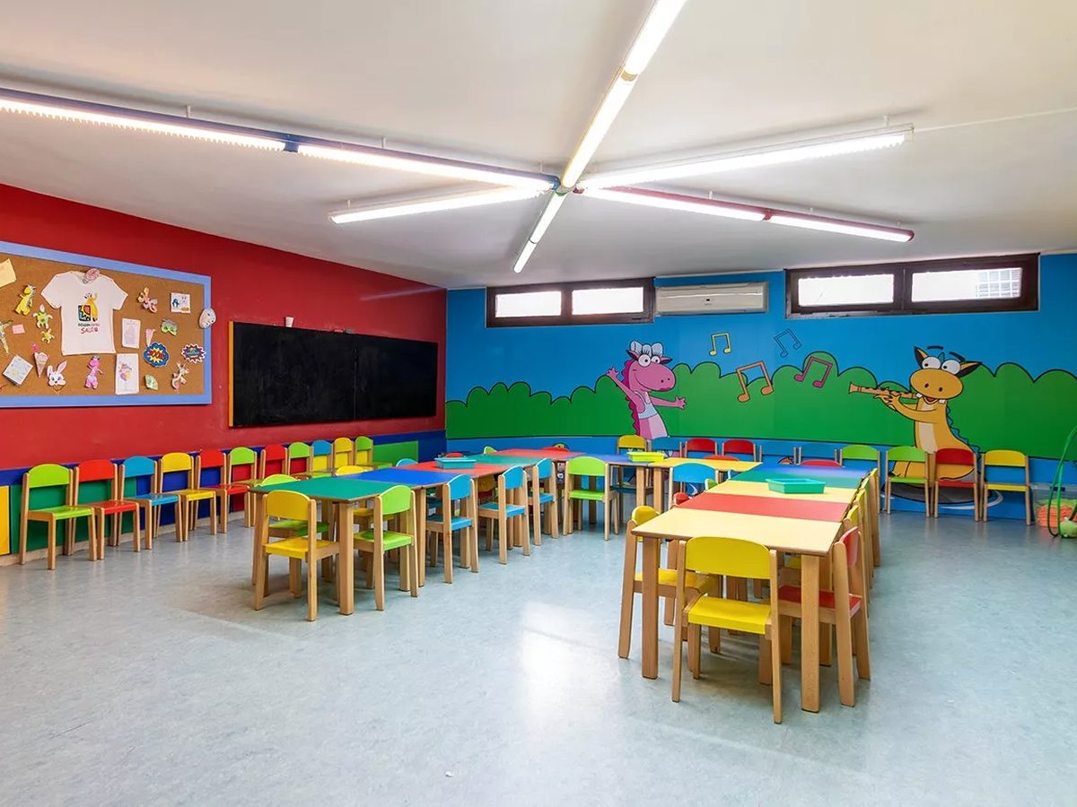 A colourful indoor kids' club room with small tables and chairs, a chalkboard, and cartoon murals on red and blue walls.