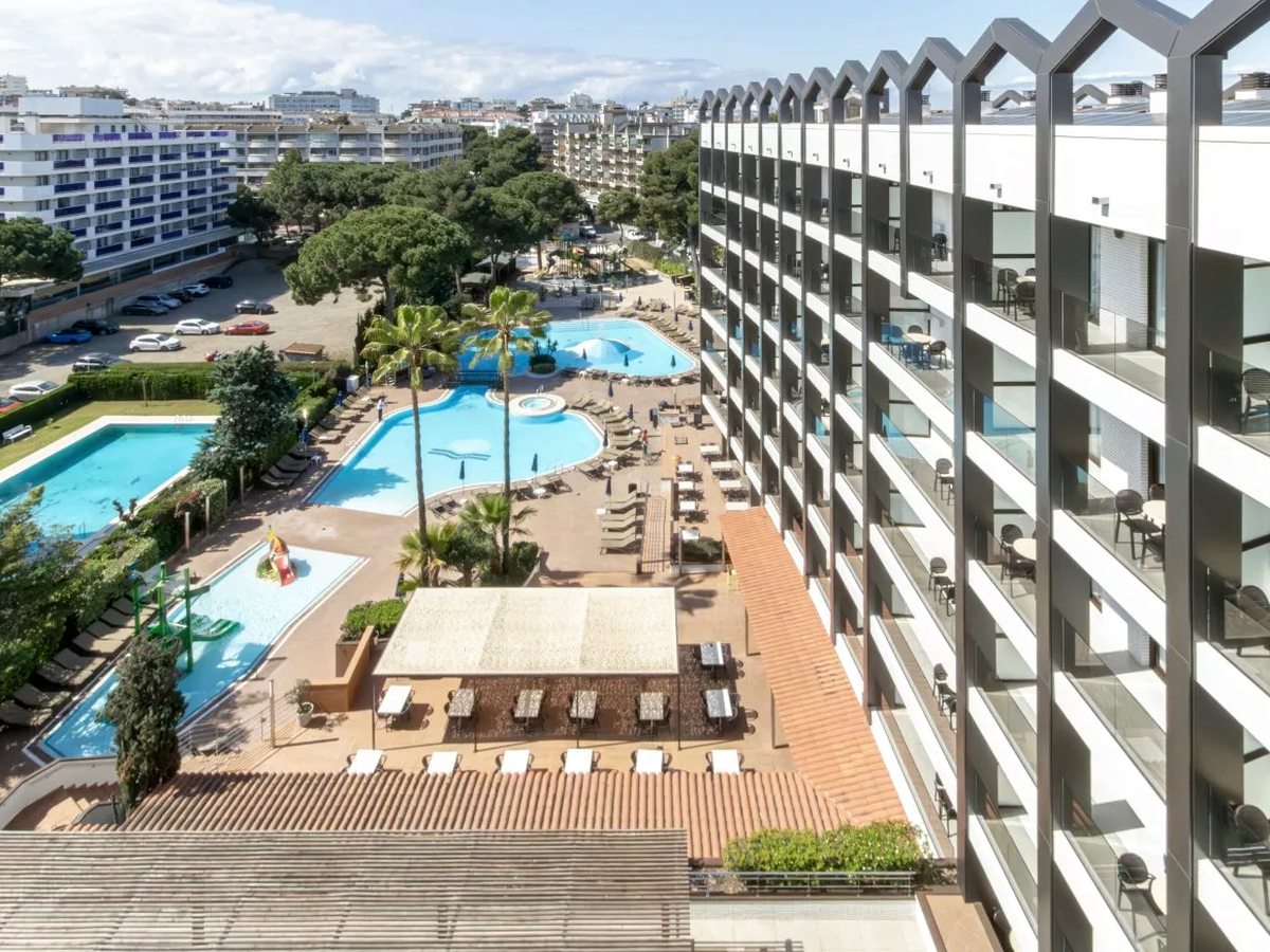 An upper-floor view of the white aparthotel block with its stepped roofline on the right, and on the left the pool courtyard with the main pool, a children's pool with slides, a rectangular pool, a covered dining terrace, palms and the town of Salou beyond.