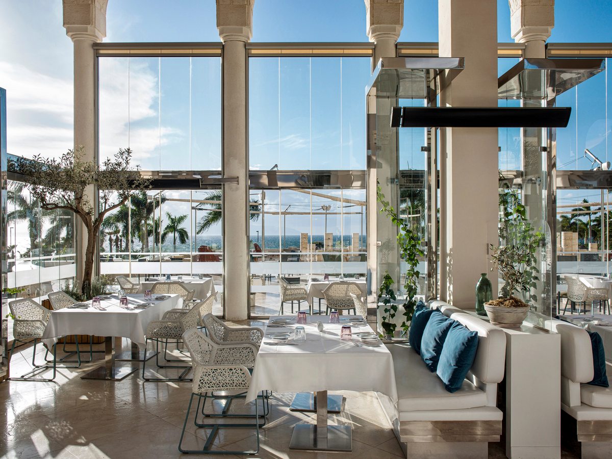 A restaurant with floor-to-ceiling windows and white-clothed tables looking out over the pools and sea.