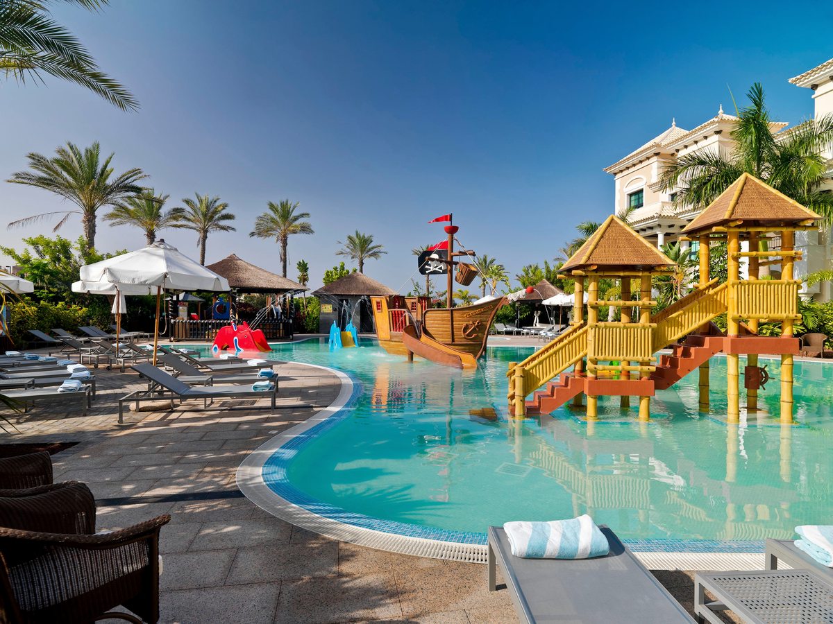 A shallow children's pool with a wooden pirate ship, a bamboo-framed slide tower and fountains, loungers around the edge.
