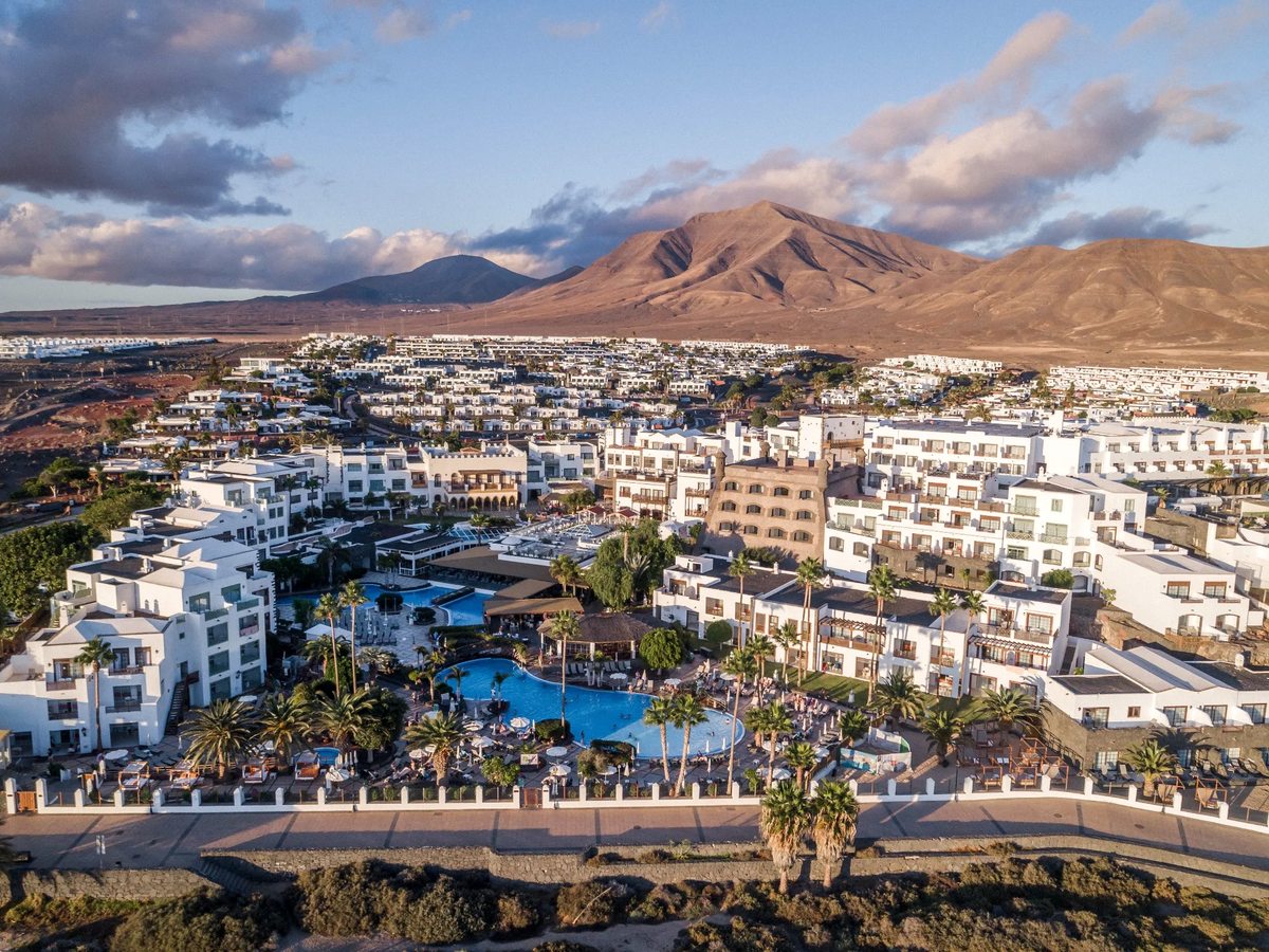 Golden-hour aerial view of white low-rise resort buildings and blue pools on a flat seafront strip, with arid volcanic mountains behind.