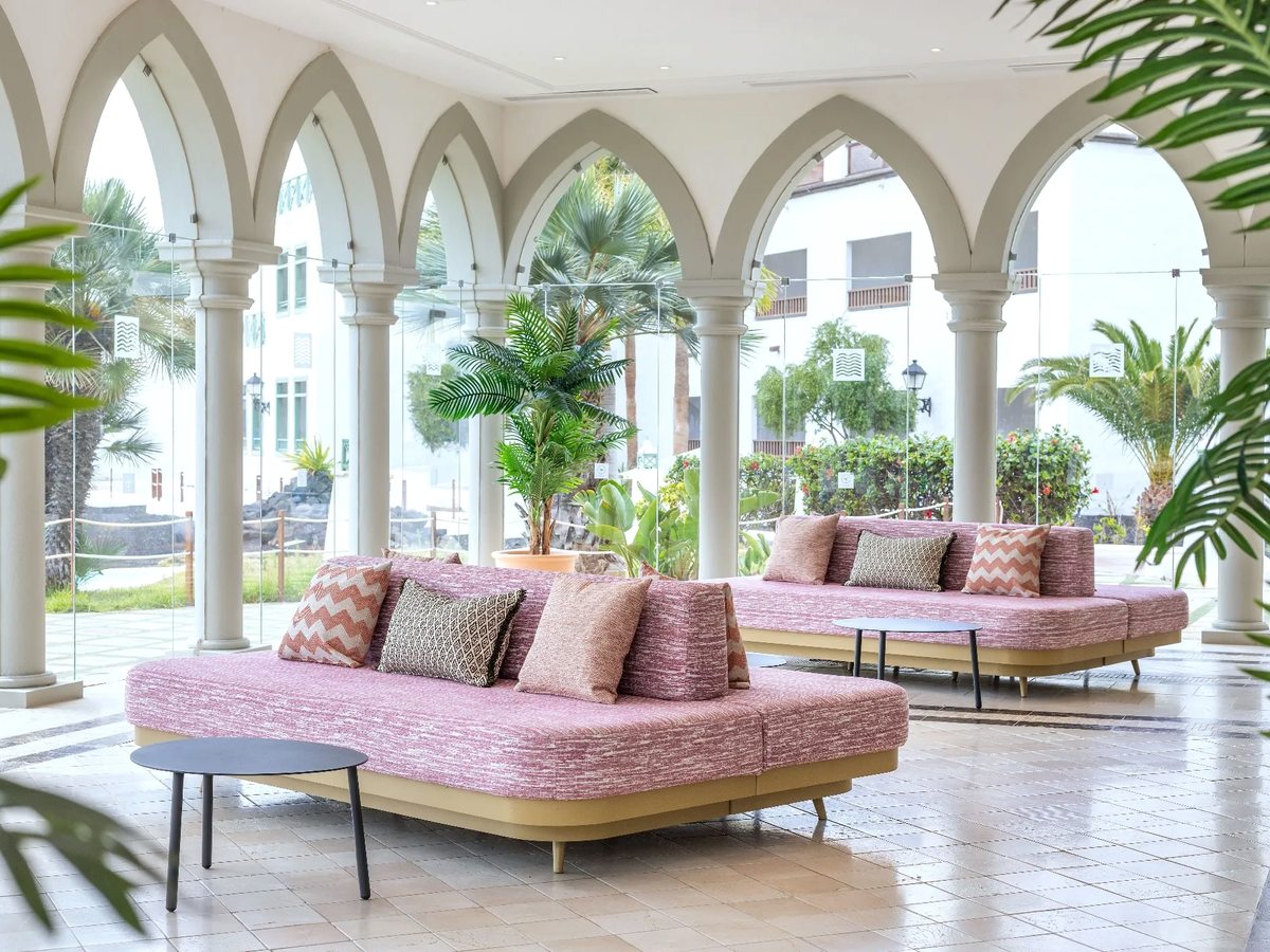 Open-sided lobby lounge with pink upholstered sofas and low tables under a white arched colonnade, opening onto a planted courtyard.