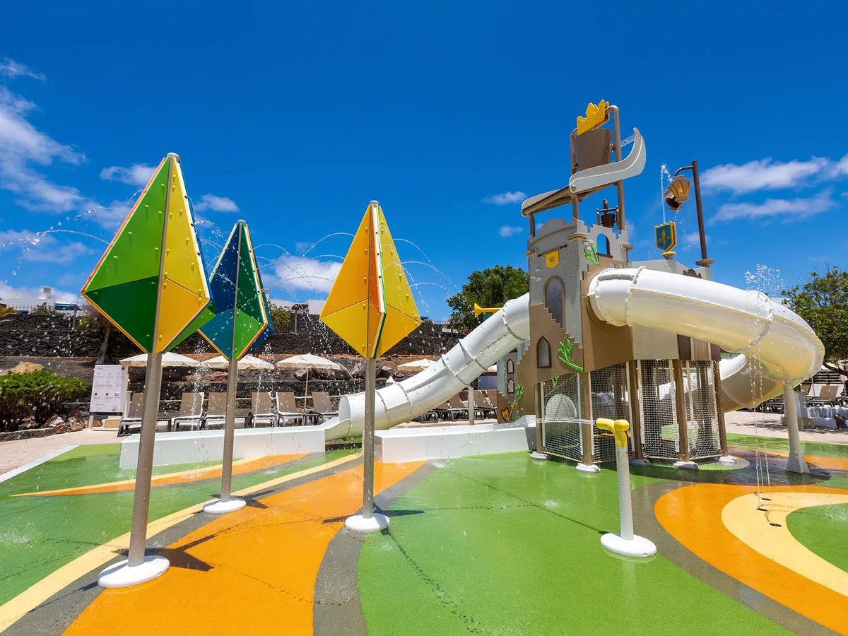Castle-themed splash park with a tower and tube slide, spray jets and colourful sail-shaped fountains on soft green-and-orange play flooring under a blue sky.
