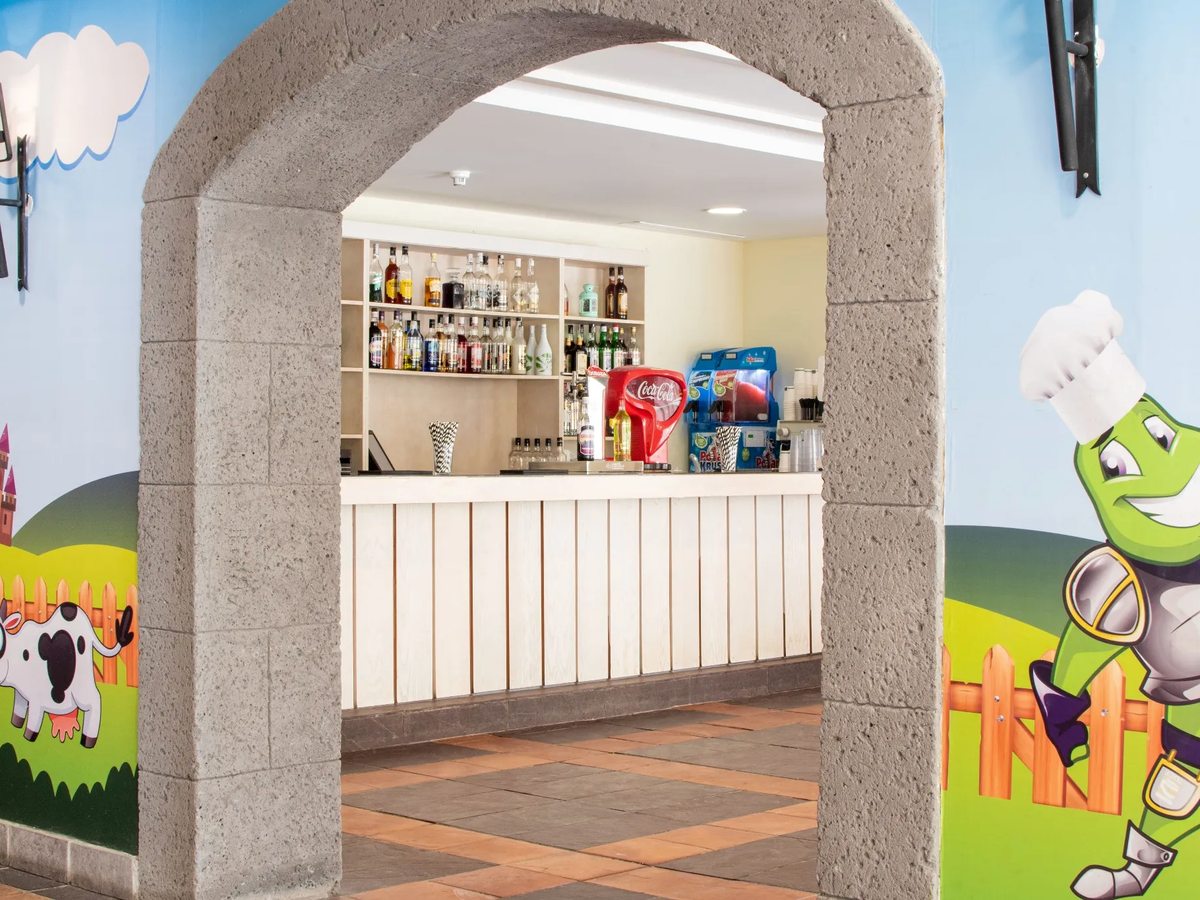 Children's snack bar seen through a stone archway, with a white counter, soft-drinks dispensers and cartoon farm and mascot murals on the walls.