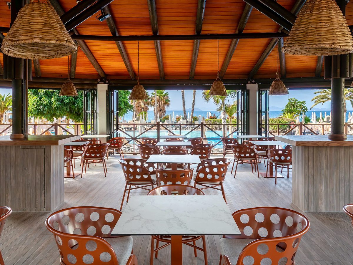Open-sided poolside restaurant with terracotta chairs, marble-topped tables and woven pendant lights under a timber ceiling, looking over the pool and sea.