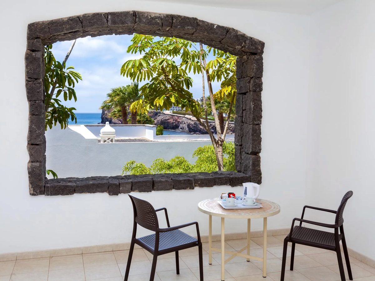 Covered room terrace with a rustic stone-arch opening, a small round table and two chairs, looking over white buildings and greenery to the sea.