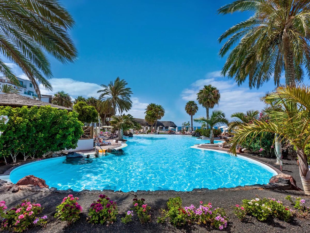 Large freeform blue pool edged with palms, flowering borders and volcanic rock, with the sea beyond and low white buildings to the side.