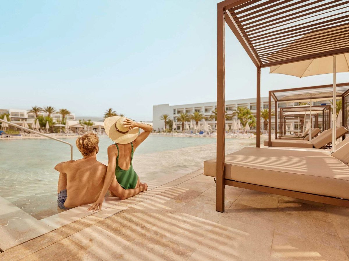 A curtained Balinese daybed under a timber canopy on the pool deck, with more daybeds and the resort beyond the water.