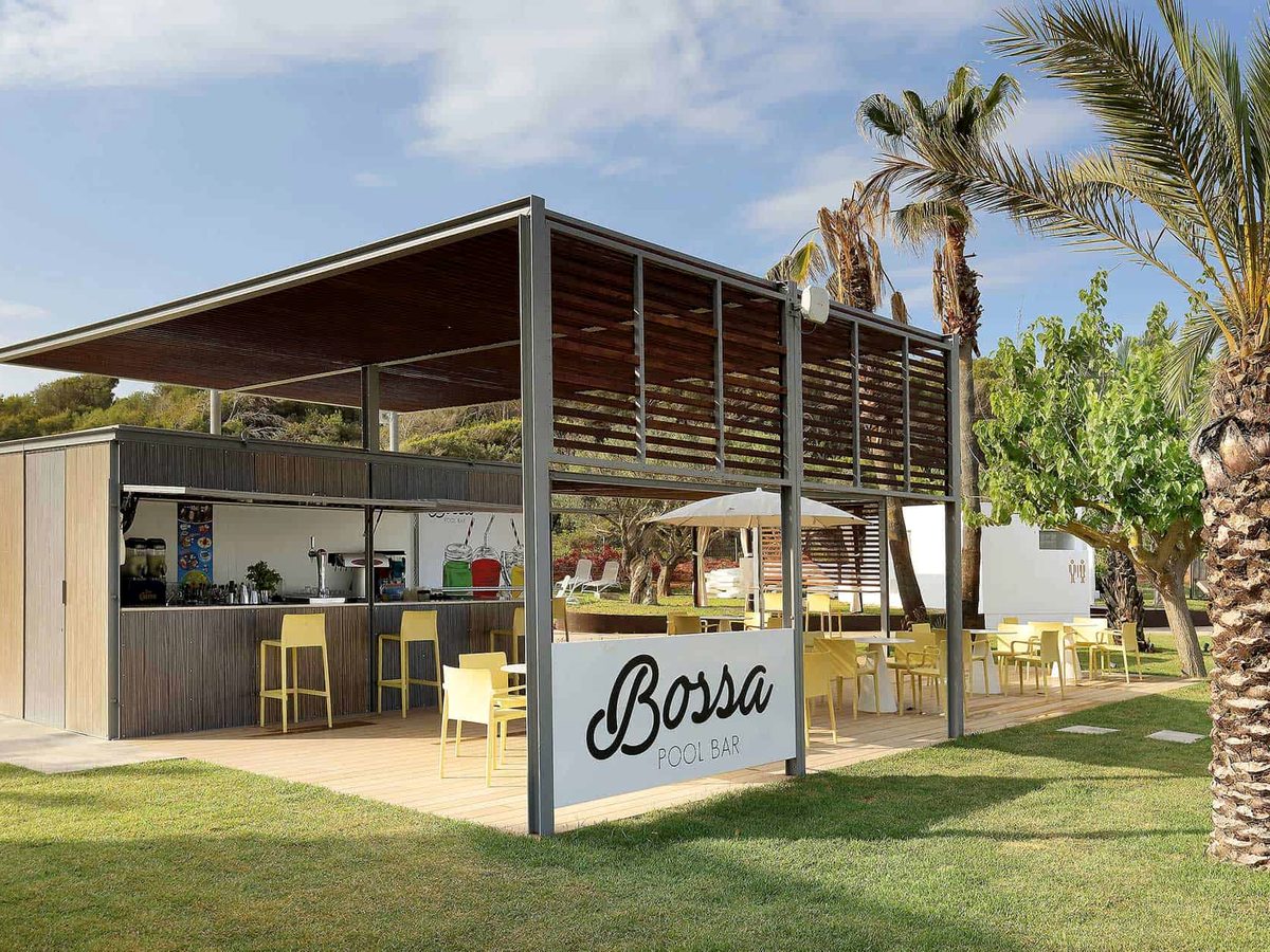 Open-sided Bossa pool bar with a timber counter, yellow bar stools and chairs under a slatted pergola on a deck beside the lawn.