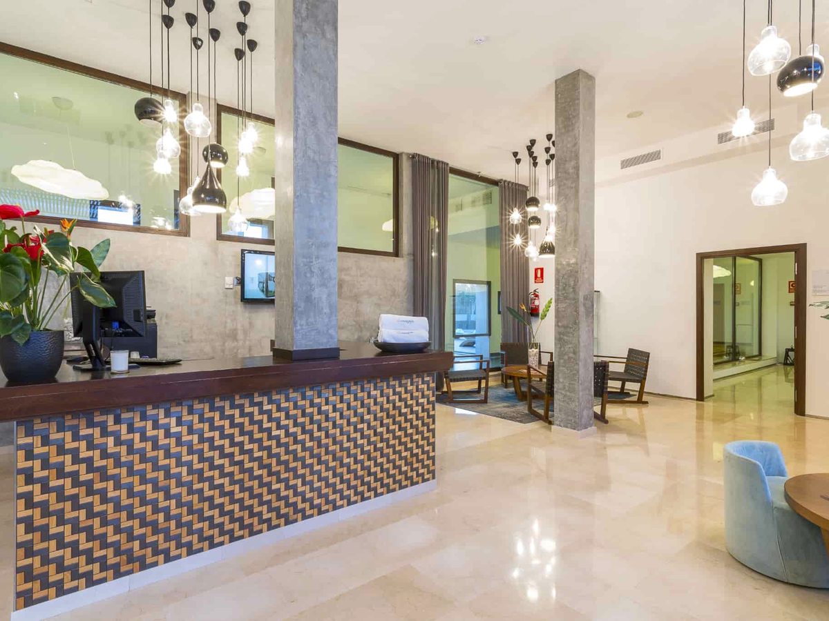 Reception area with a patterned front desk, pendant lights and wall-mounted screens between concrete columns over a polished floor.