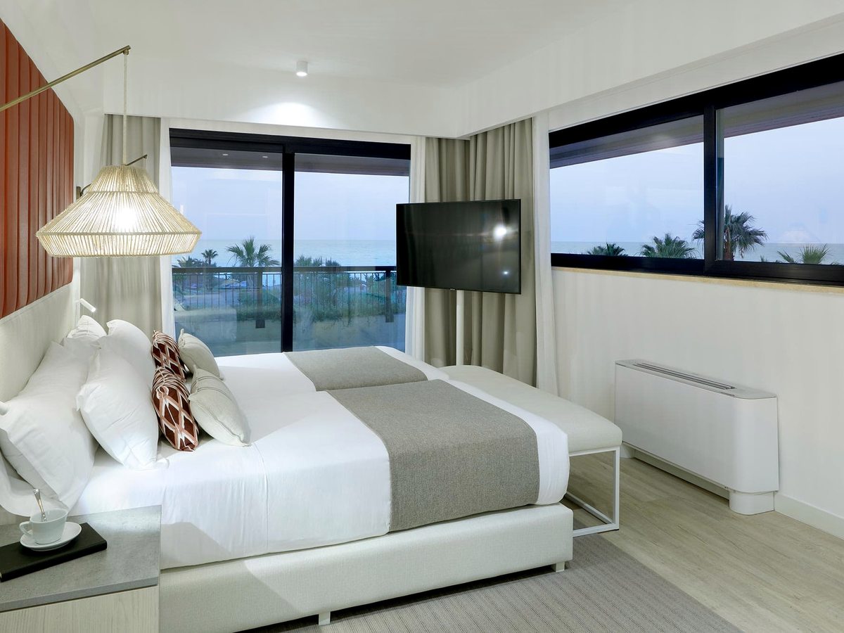 A room with a king bed, a bench at its foot and a wall-mounted TV, with large windows and a balcony looking out to the sea.