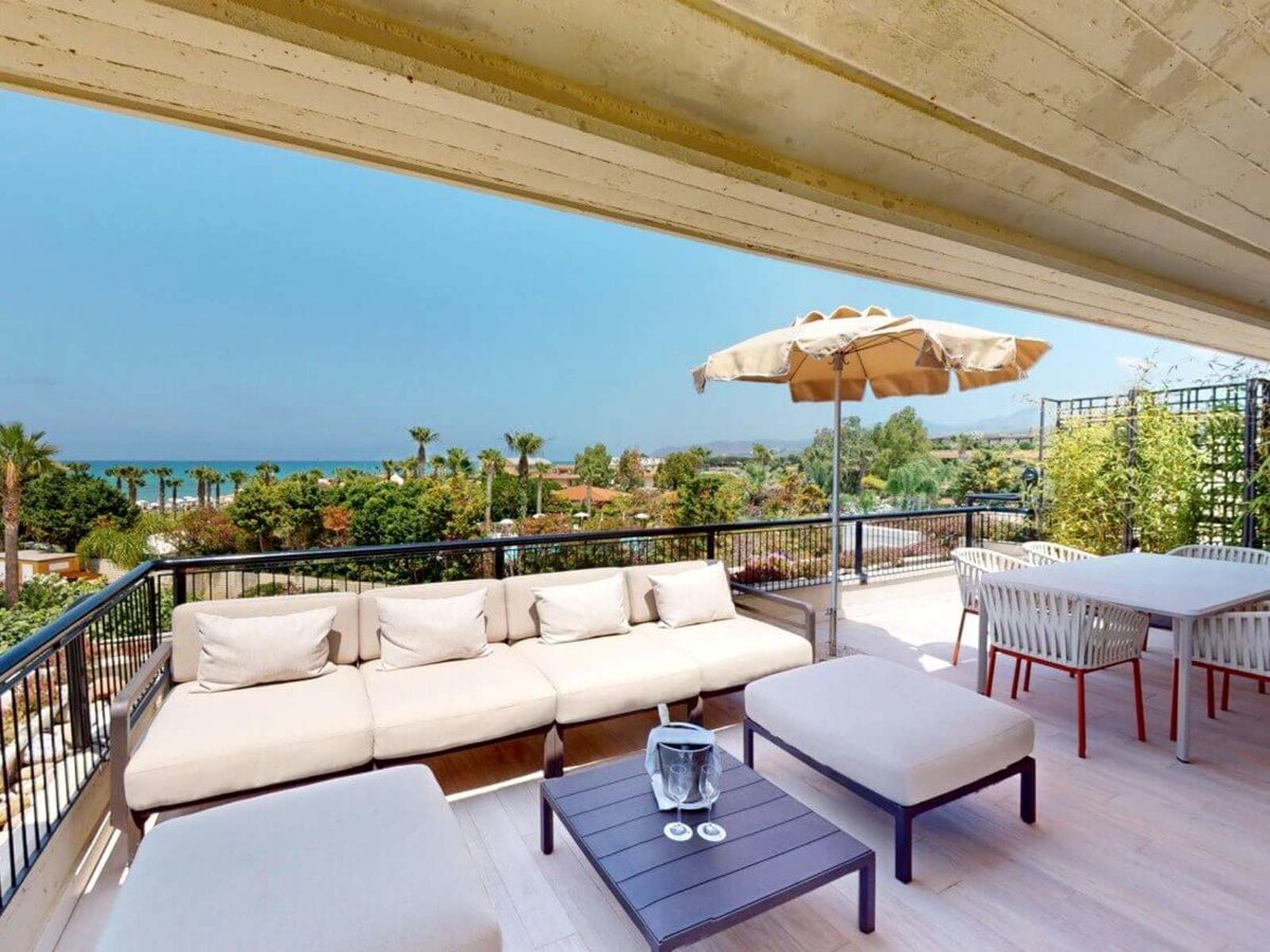 A wide covered terrace with a long cream sofa, an ottoman, a parasol and a dining table, looking out over the resort's palm gardens to the sea and the hills behind.