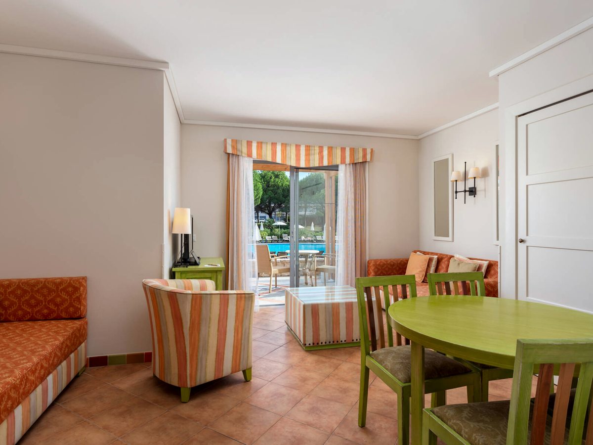 An open-plan villa living room with orange sofas, a green dining table and patio doors to a pool terrace.