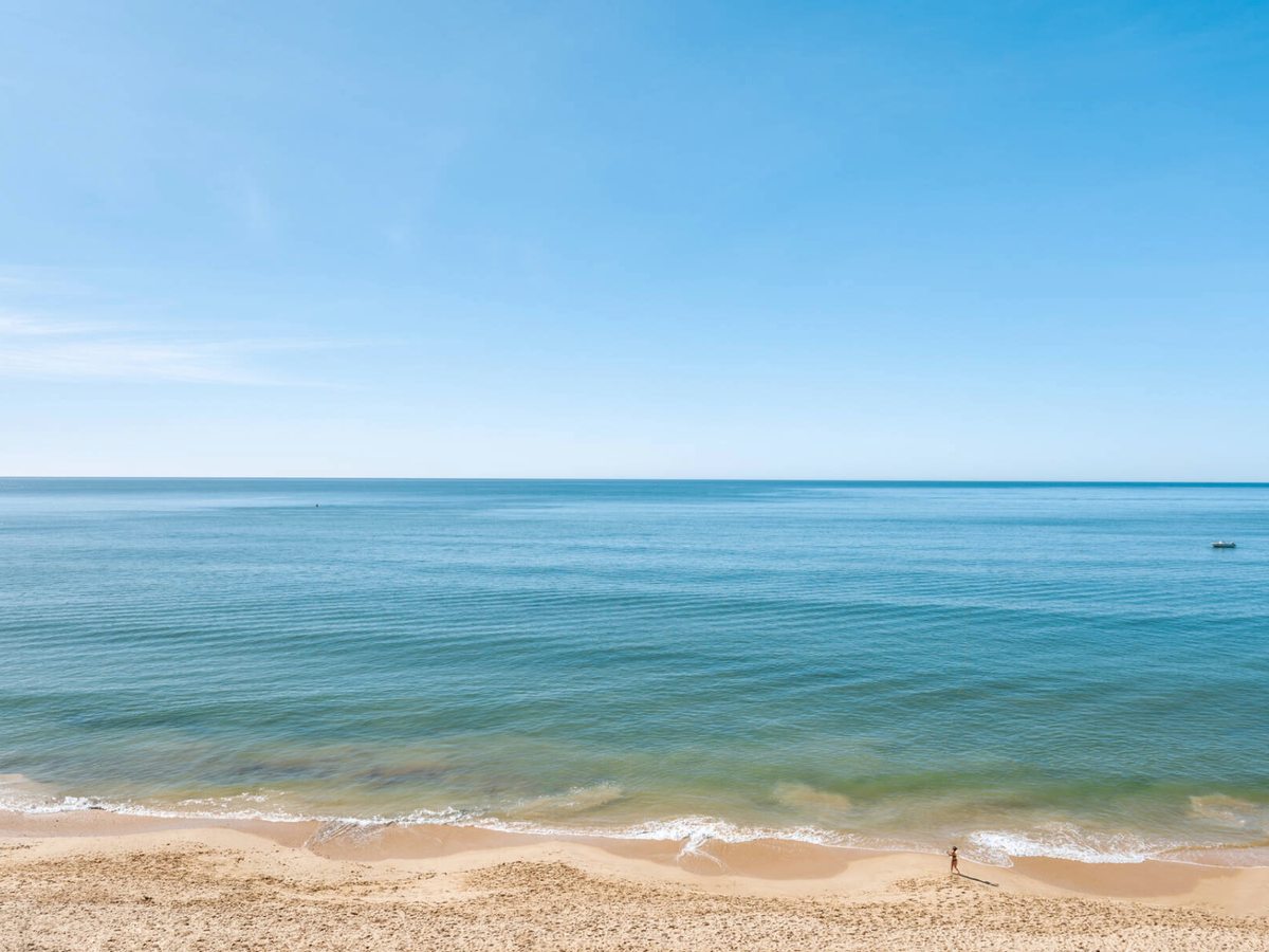A wide sandy beach meeting calm turquoise water, with a single figure at the shoreline.
