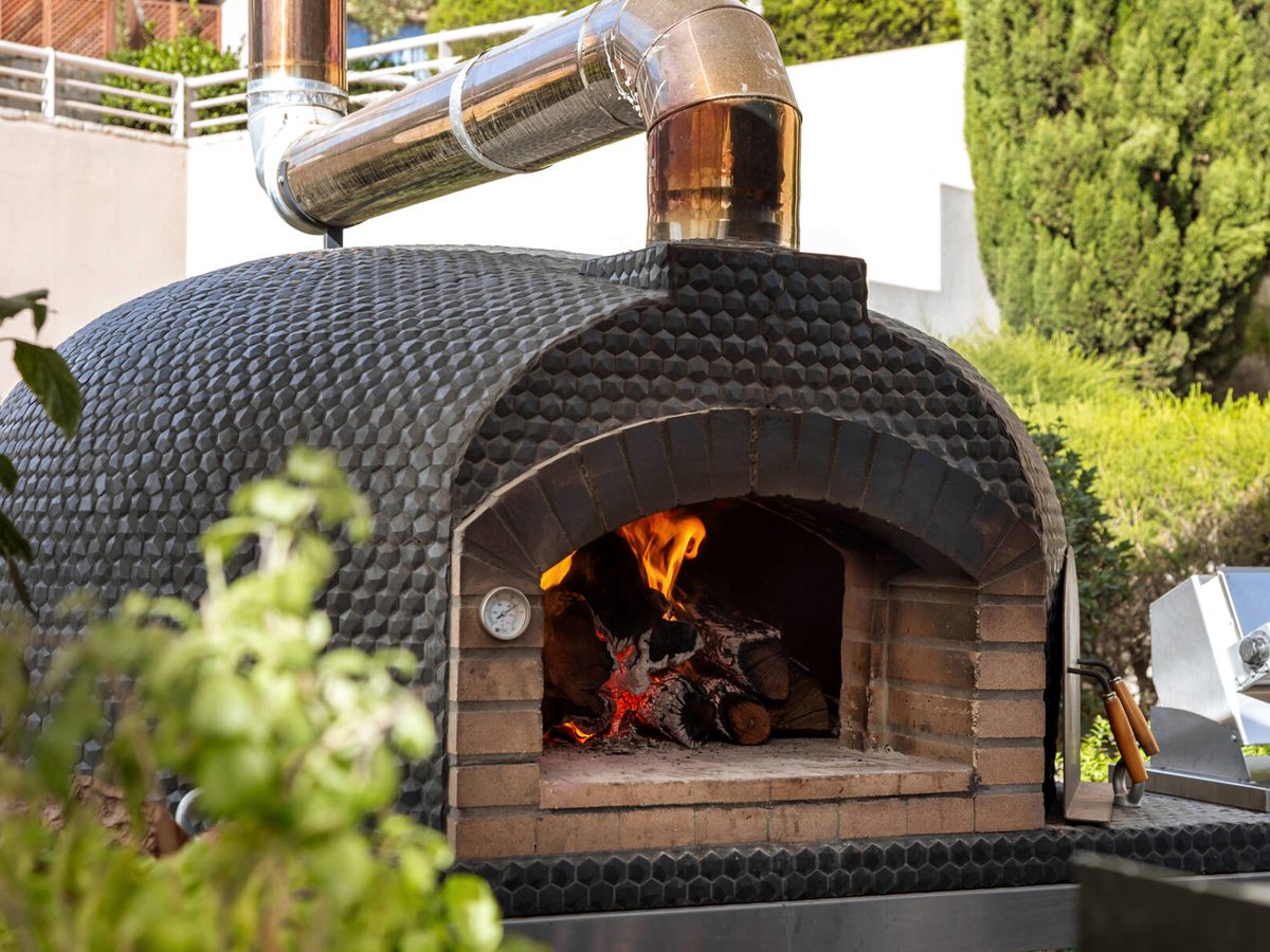 A black-tiled wood-fired pizza oven with a lit fire and a steel flue, set in a garden.