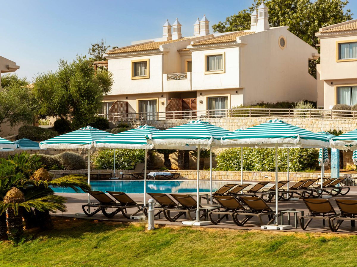 A pool with green striped parasols and loungers on a lawn, villas rising behind.