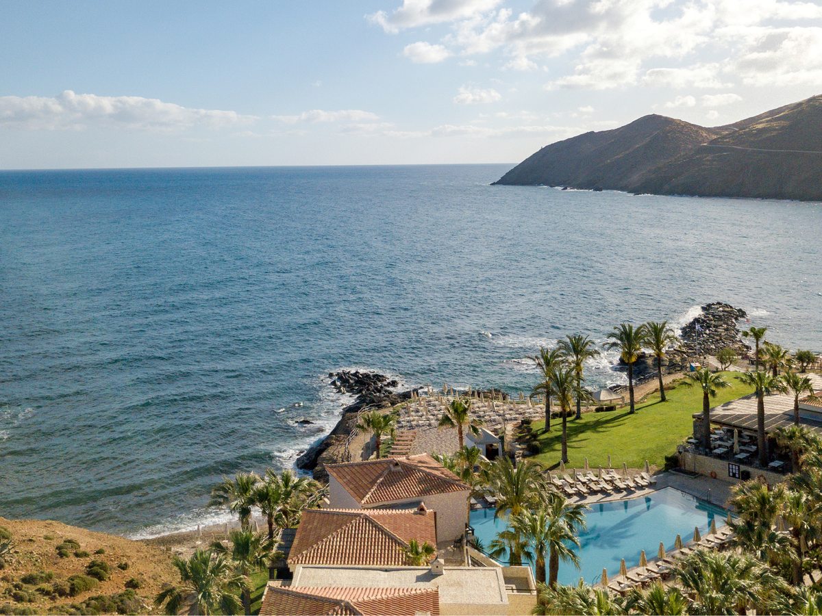 Elevated view over the resort's low tile-roofed buildings, pool and gardens on a sandy bay, with the sea and a rocky headland beyond.