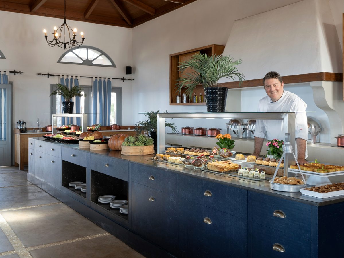 Long buffet counter with hot and cold dishes and a chef behind it, under a timber ceiling.