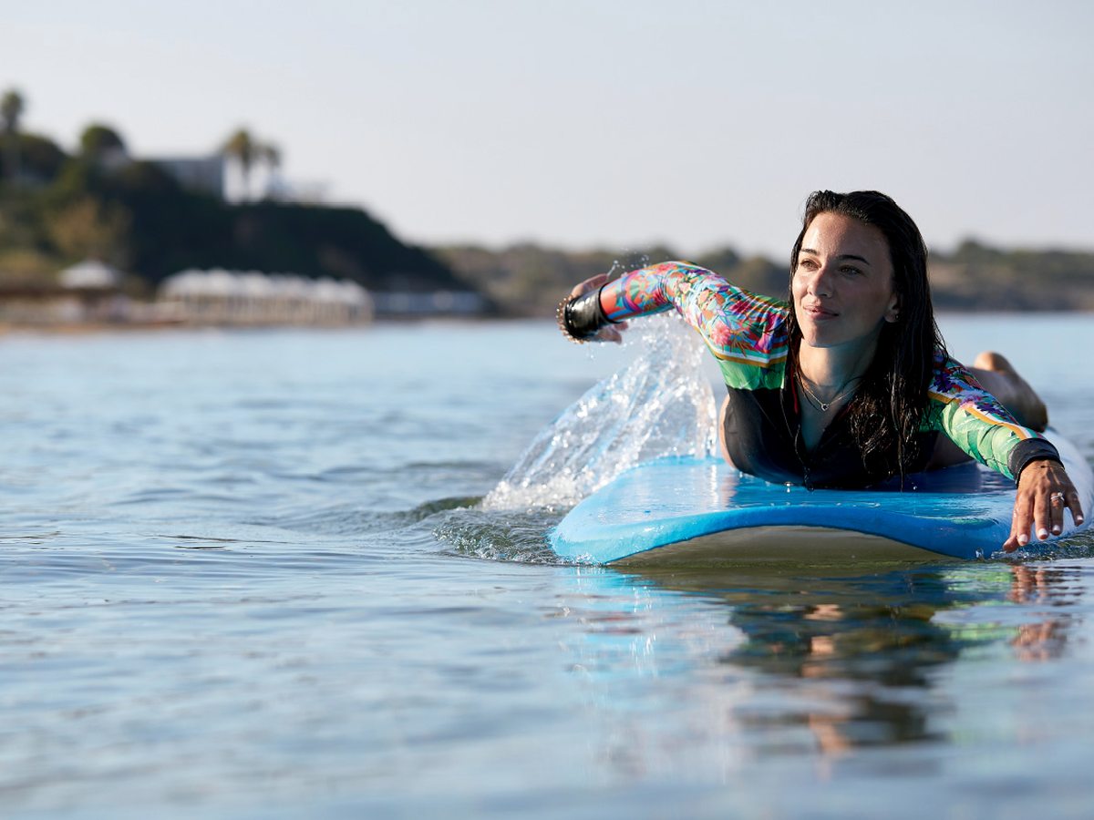 A woman lying prone on a foam board, hand-paddling in calm shallow water off the beach.