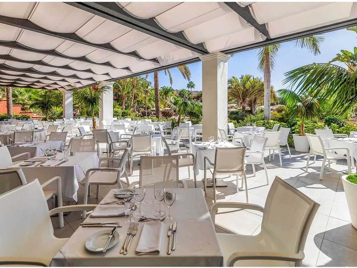 A covered outdoor restaurant terrace set with white chairs and marble-topped tables among potted plants.