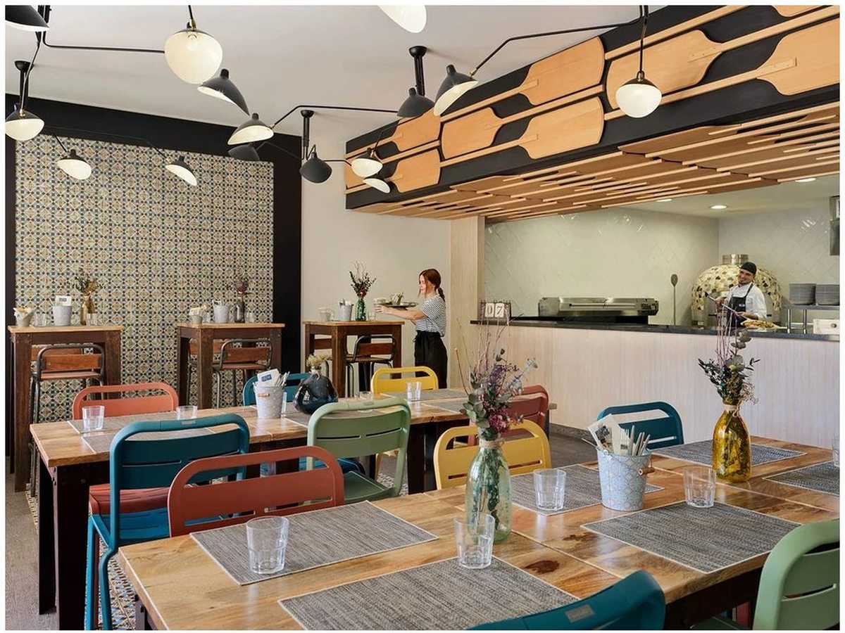 An indoor dining room with colourful mismatched chairs at wooden tables, pendant lights and a service counter with staff along the far wall.