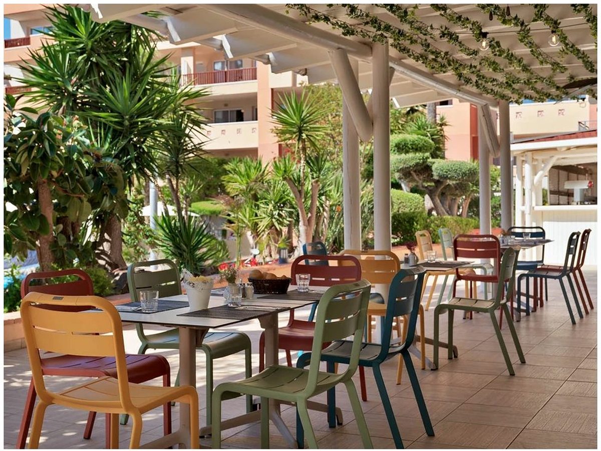 A covered outdoor restaurant terrace with colourful chairs at small tables, potted greenery and palm trees, with apartment balconies behind.